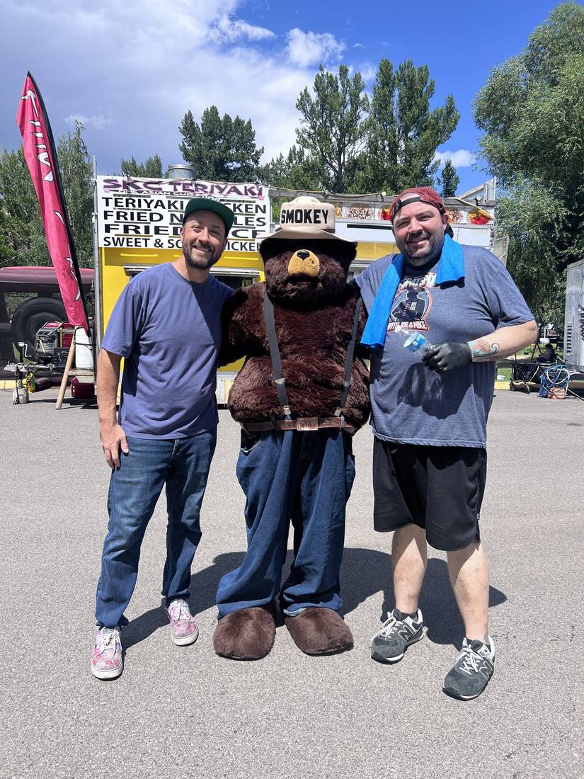 Two House of Hogs teammates posing with Smokey Bear in front of a neighboring food truck